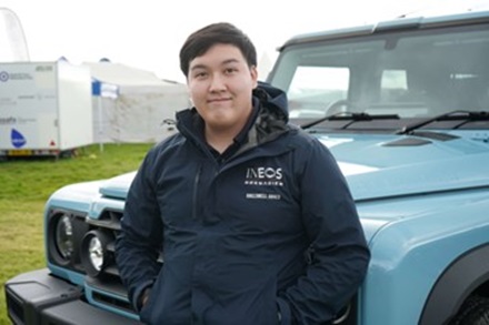 A male student standing outdoors next to large motor vehicle.