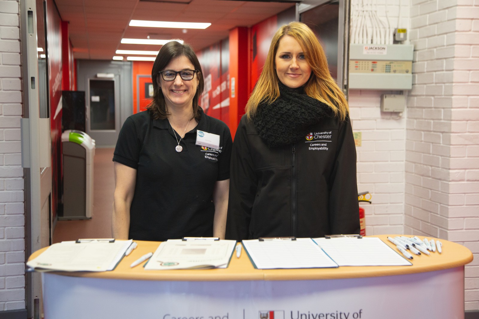 Two members of staff from Careers Service standing behind a desk with papers and pens, facing the camera, smiling.