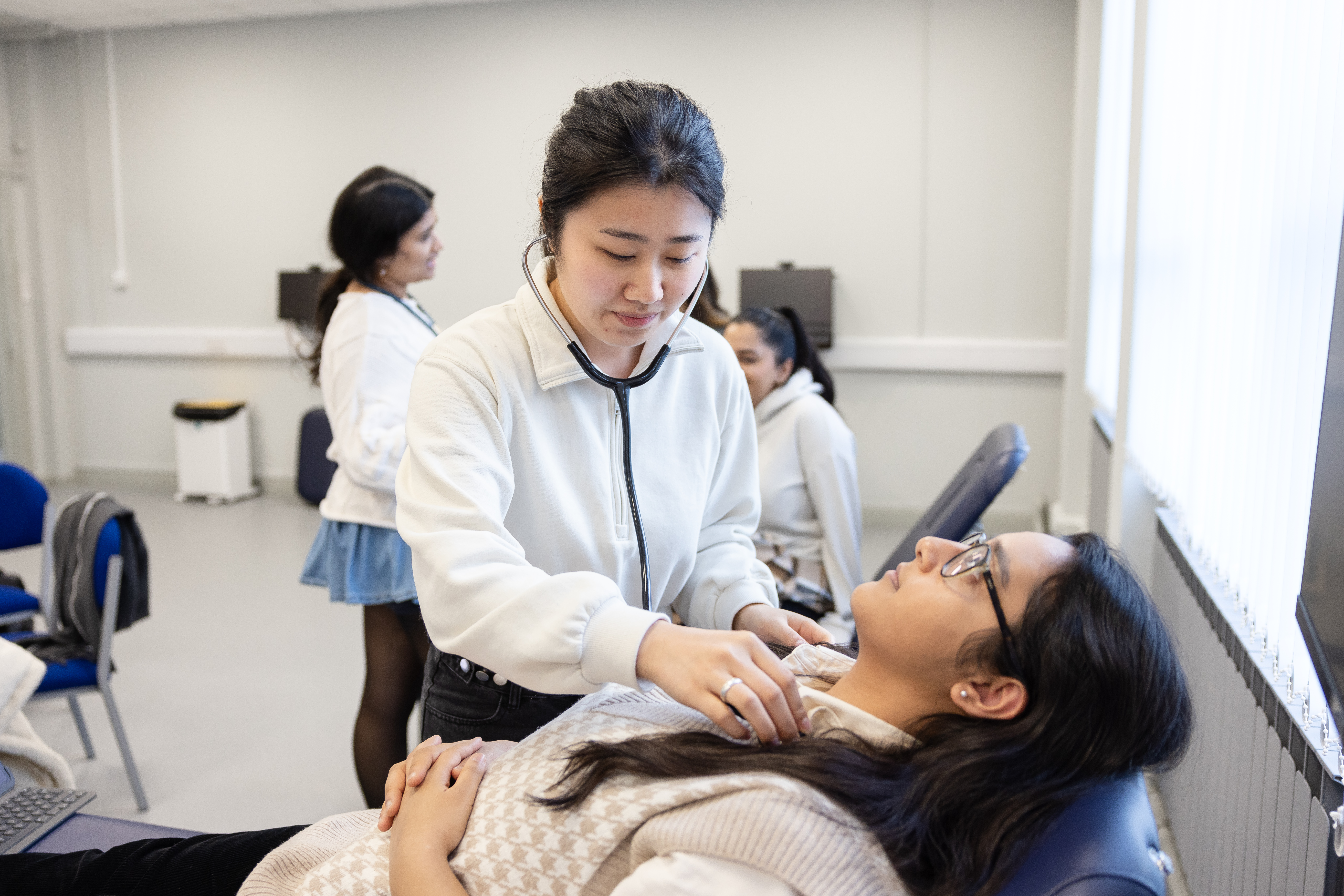 A young female student using a stethoscope on another student.