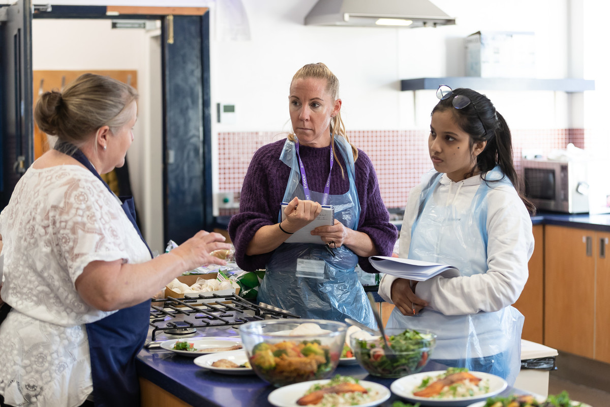 Three people talking in a kitchen wearing aprons and gloves with a plates of food on a worktop.
