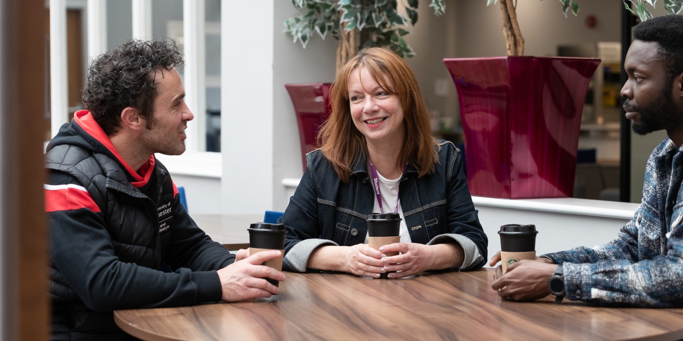 Two students and a member of the Residential Services team are siting around a table, outside having a coffee. One student faces the camera, the other student is facing the camera from the side, as is the Residential Advisor but from the opposing side.